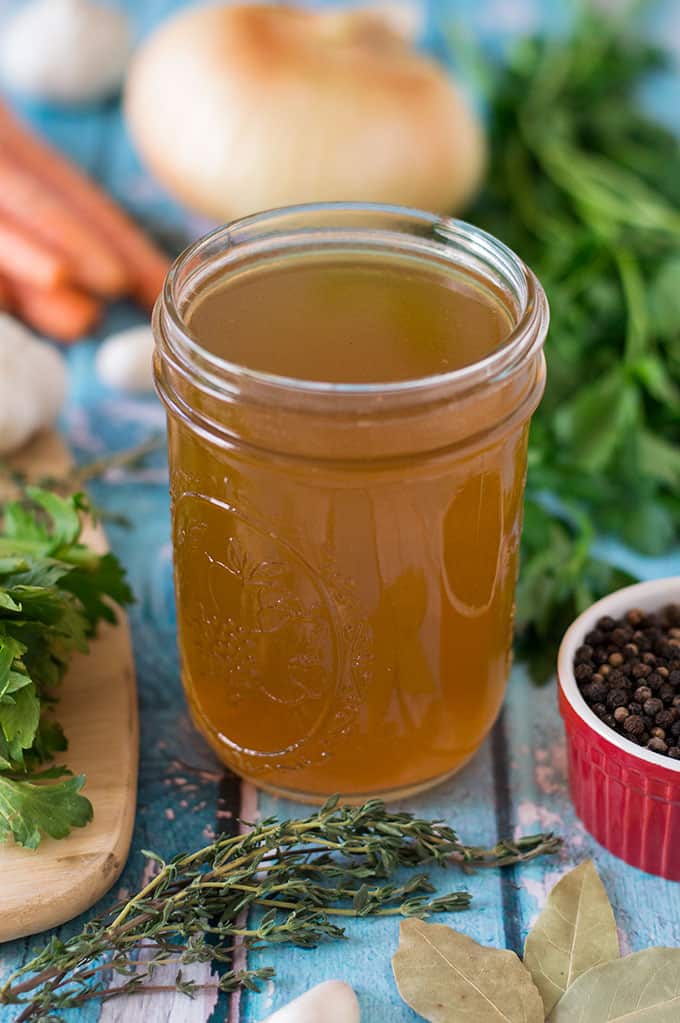 A jar of chicken stock surrounded by celery, carrots, onions, garlic, herbs, and spices.