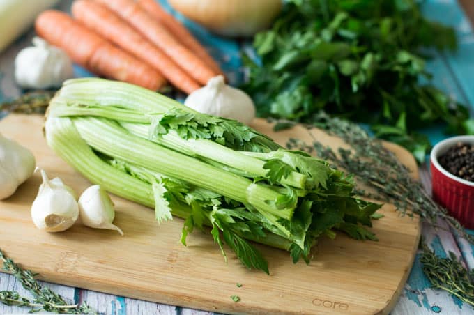 Celery, garlic cloves, herbs, carrots, and onions, on a tabletop.