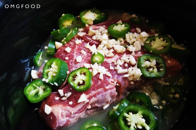 A closeup of a raw beef roast, sliced jalapeños, and chopped garlic in a pot.