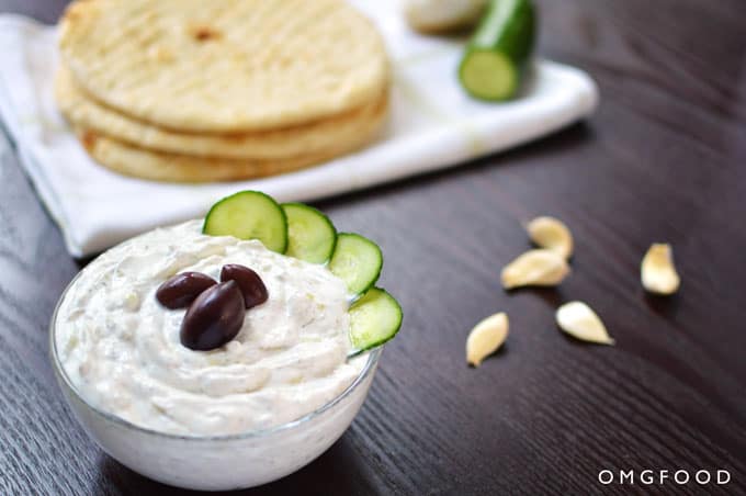 A bowl of tzatziki with pita bread and garlic cloves in the background.