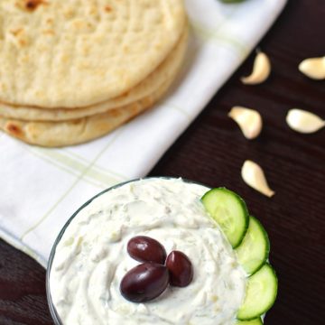 A bowl of tzatziki dip with pita bread and garlic cloves in the background.