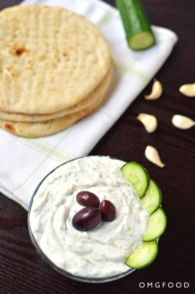 A bowl of tzatziki dip with pita bread and garlic cloves in the background.