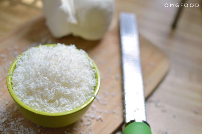 Closeup of grated cheese in a bowl next to a grater and ball of cheese.