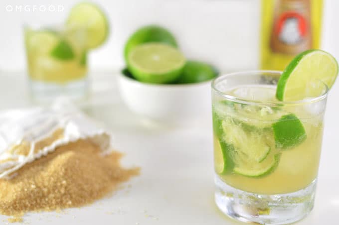 Close up of a cocktail in a drinking glass, bag of sugar, and bowl of limes on a tabletop.