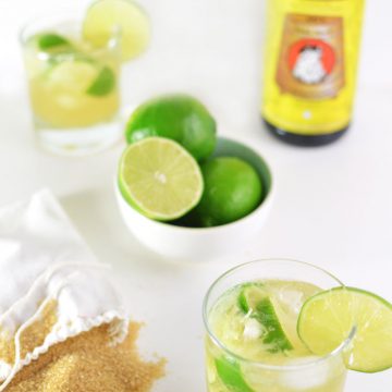 A bowl of limes, bag of sugar, and cocktails on a tabletop.