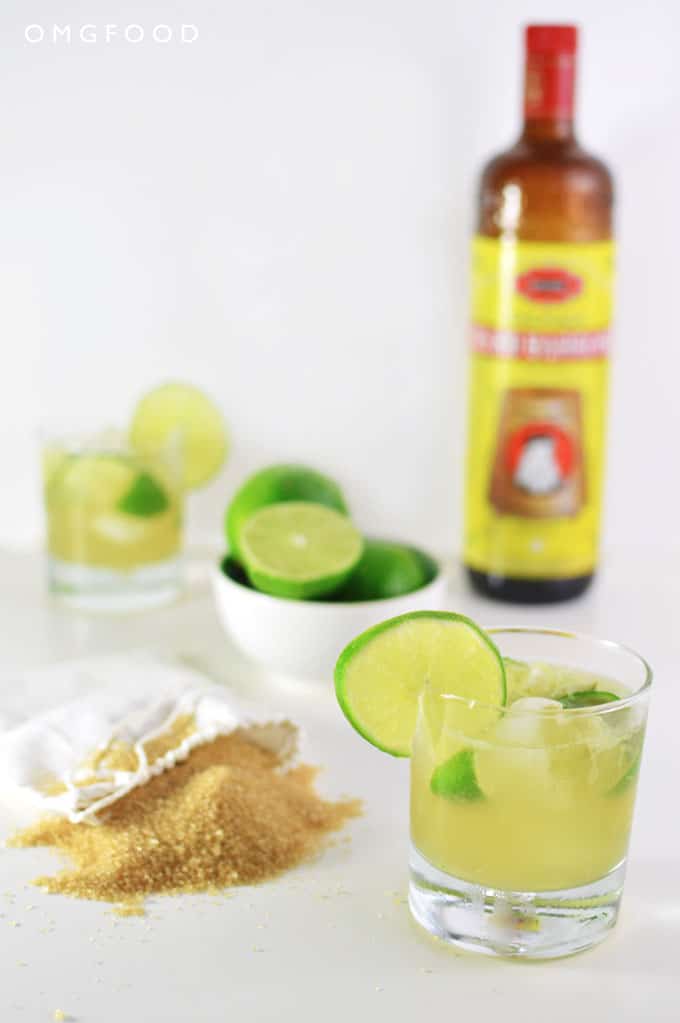 A cocktail in a drinking glass, bag of sugar, and bowl of limes on a tabletop.