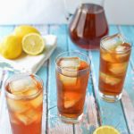 Three tall glassed of iced tea with lemons and a pitcher of iced tea in the background.