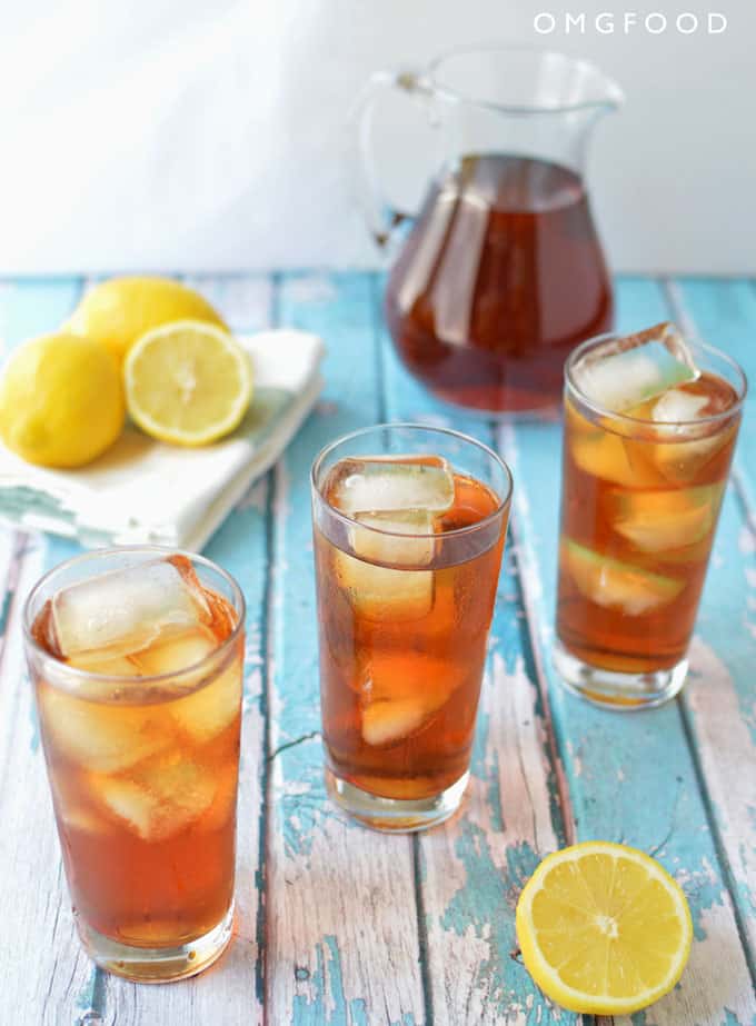 Three tall glassed of iced tea with lemons and a pitcher of iced tea in the background.