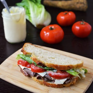 A sandwich on a cutting board with tomatoes, lettuce, and a jar of mayo in the background.