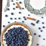 A blueberry pie on a tabletop with forks, a plate, and scattered blueberries.