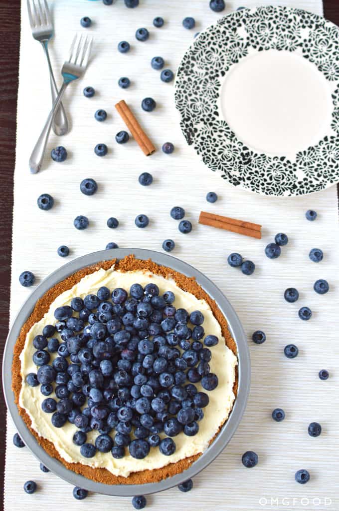 A blueberry pie on a tabletop with forks, a plate, and scattered blueberries.