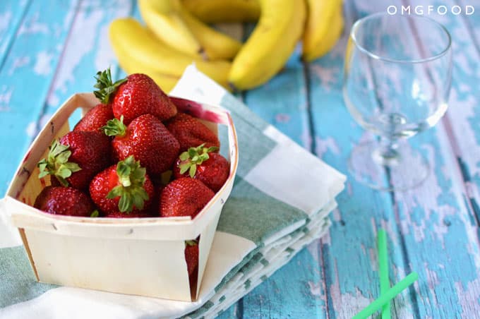 A box of strawberries on a tabletop with bananas in the background.