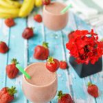 Two strawberry smoothies in glasses on a tabletop with strawberries and small vase of red flowers.