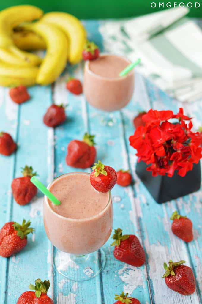 Two strawberry smoothies in glasses on a tabletop with strawberries and small vase of red flowers.