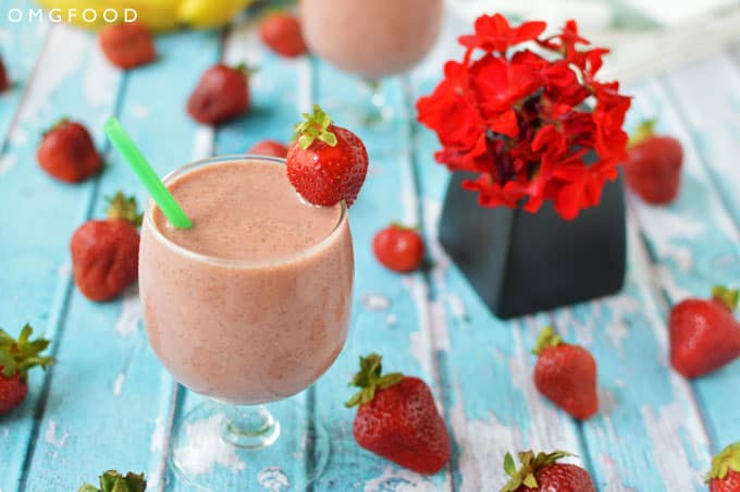 Strawberry smoothie in a drinking glass on a tabletop with strawberries and a small vase of red flowers.