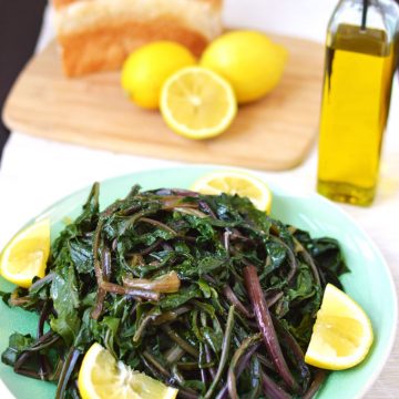 A plate of cooked dandelion greens with olive oil, bread, and lemons in the background.