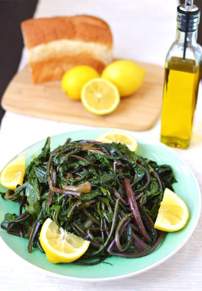 A plate of cooked dandelion greens with olive oil, bread, and lemons in the background.