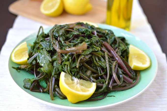 Closeup of a plate of cooked dandelion greens with olive oil, bread, and lemons in the background.