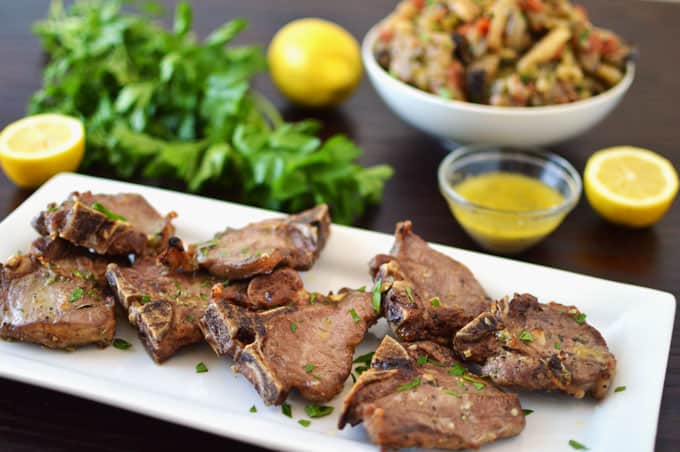 A plate of lamb loin chops with parsley, lemon, and vegetables in the background.