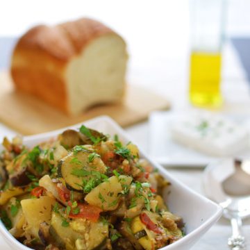 A bowl of baked vegetables with bread and olive oil in the background.