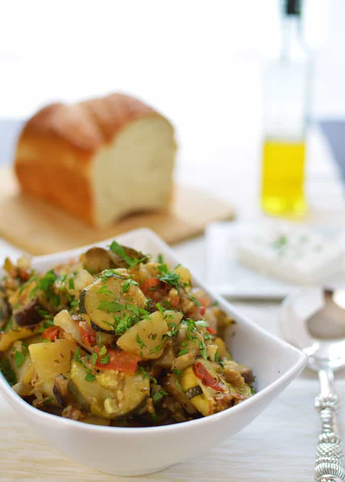 A bowl of baked vegetables with bread and olive oil in the background.