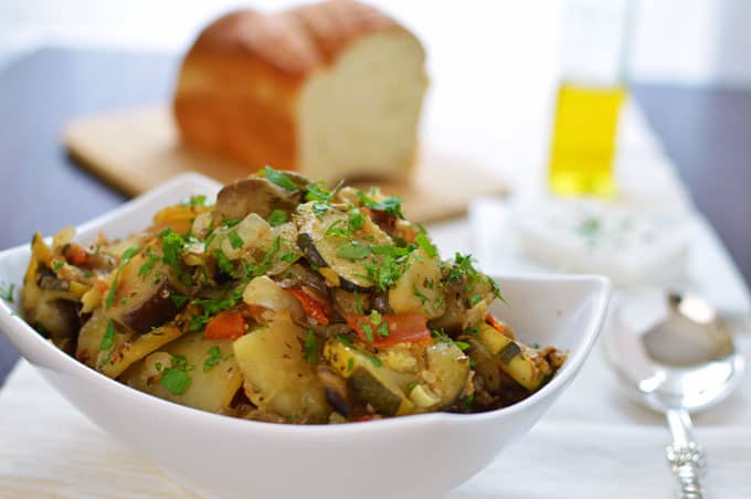 A bowl of baked vegetables with bread and olive oil in the background.