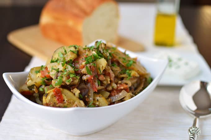 Closeup of a bowl of baked vegetables with bread and olive oil in the background.