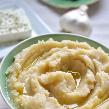 Whipped potato dip in a bowl with garlic and feta in the background.