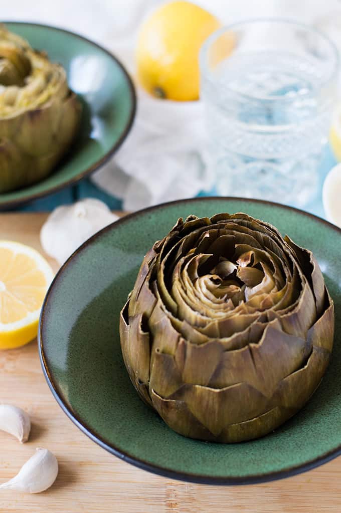 A roasted artichoke in a bowl with lemons, garlic, and glass of water on a tabletop.