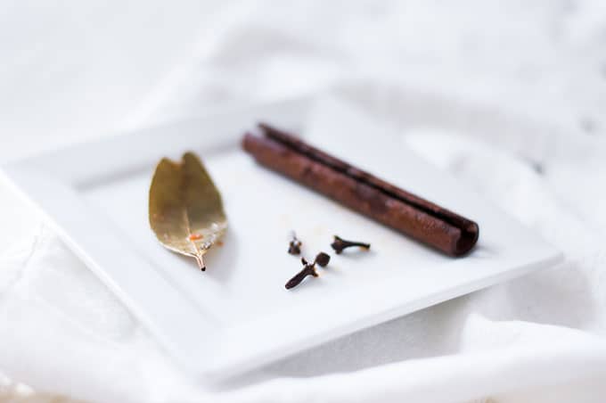 A cinnamon stick, bay leaf, and whole cloves on a small plate.
