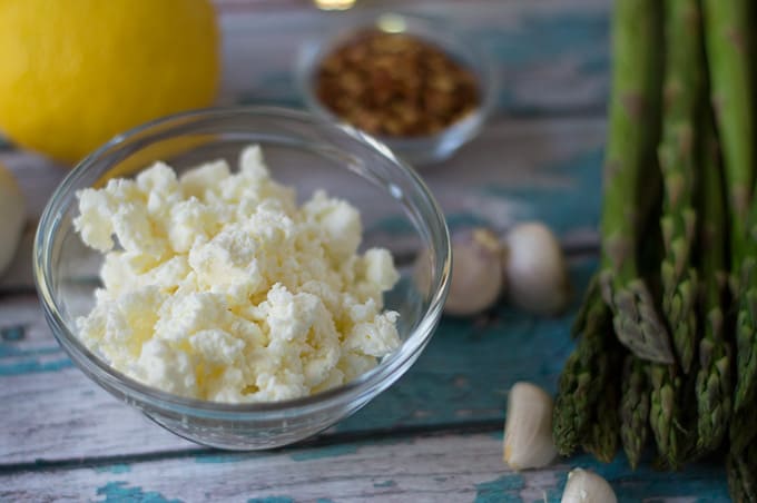 Closeup of crumbled feta in a small bowl next to garlic cloves and asparagus.