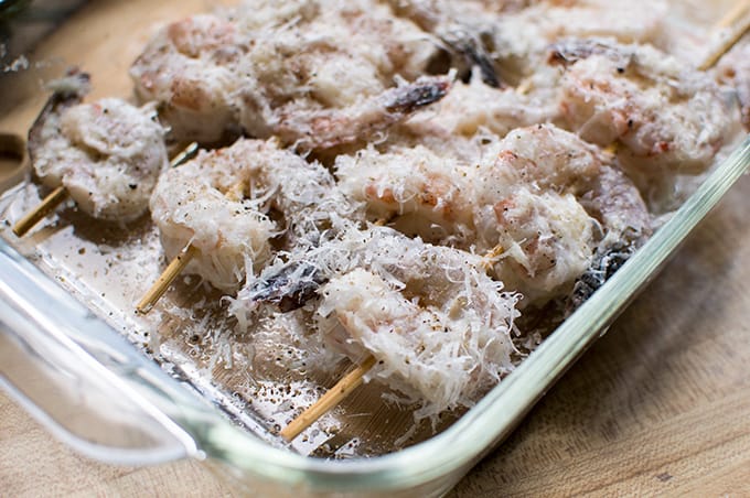 Skewered raw shrimp coated with grated cheese in a dish.