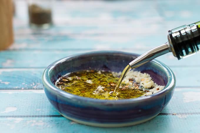 Olive oil being poured into a small dish of grated cheese, garlic, and spices.