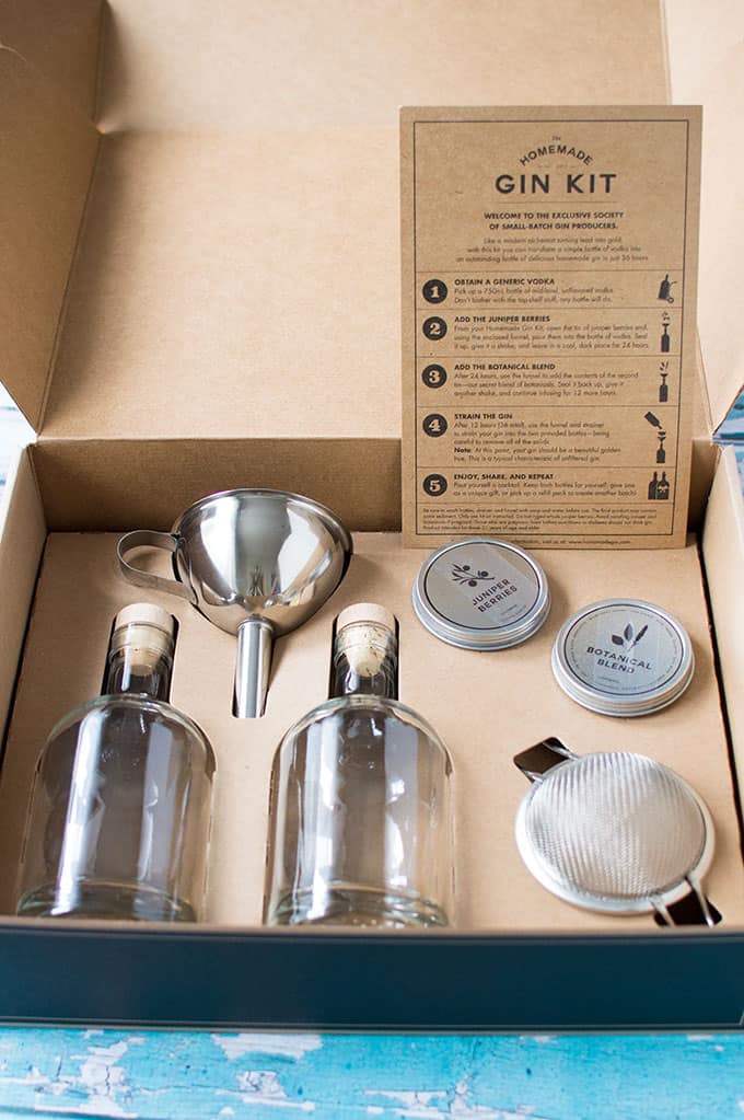 A closeup of a gin-making kit in a box, including glass bottles, a funnel, fine mesh sieve, and spices.