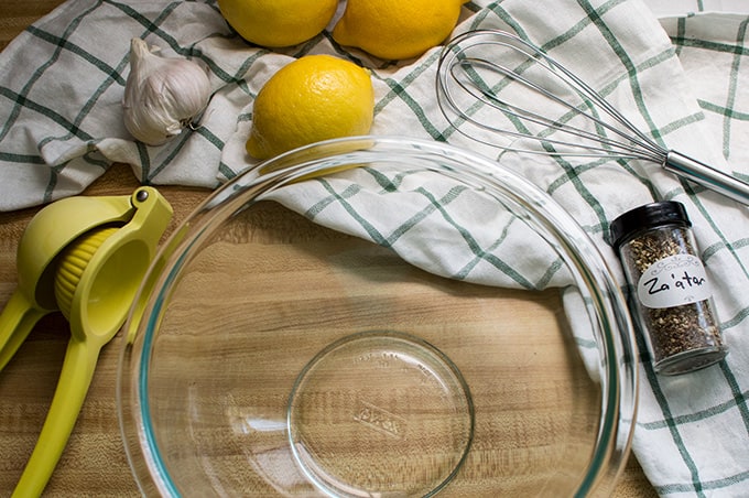 Citrus squeezer, whole lemons, a whisk, bottle of Za\'atar seasoning, and an empty glass bowl on a countertop.