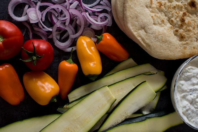 Sliced zucchini, tinkerbell peppers, sliced red onions, and stacked pita bread on a tabletop.