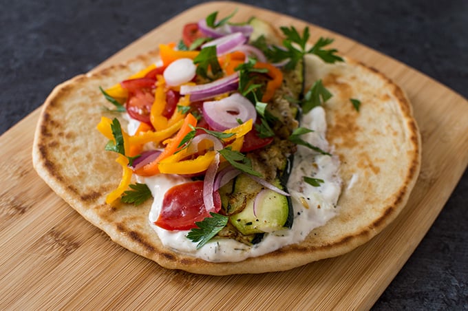 An open-faced pita wrap on a wooden cutting board topped with onions, zucchini, peppers, and tzatziki sauce.