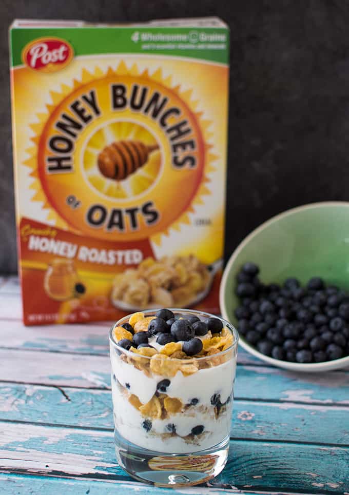 A bowl of blueberry yogurt parfait on a table with a box of cereal and bowl of blueberries.