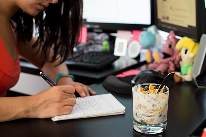 A person sitting at a desk in front of a computer while writing in a notebook.