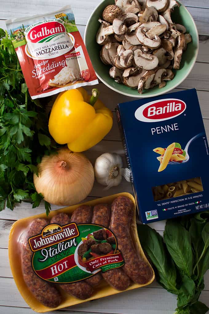 Ingredients to male one-pot creamy sausage and pasta: package of mozzarella cheese, bowl of sliced mushrooms, yellow bell pepper, whole yellow onion, bulb of garlic, box of penne pasta, package of Italian hot sausage, and fresh basil leaves.