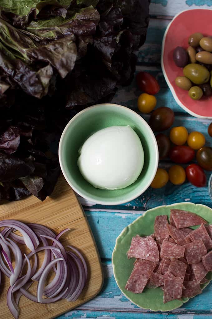 Red leaf lettuce, buffalo mozzarella, grape heirloom tomatoes, mixed olives, sliced salami and sliced onions on a table.