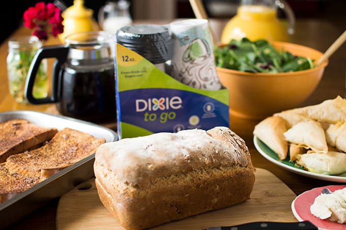 Baked French toast, a loaf of bread, a package of disposable coffee cups, a pot of coffee, salad, and juice on a table.