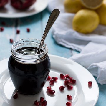 Glass jar of pomegranate molasses with a spoon inside and lemons and pomegranates in the background.