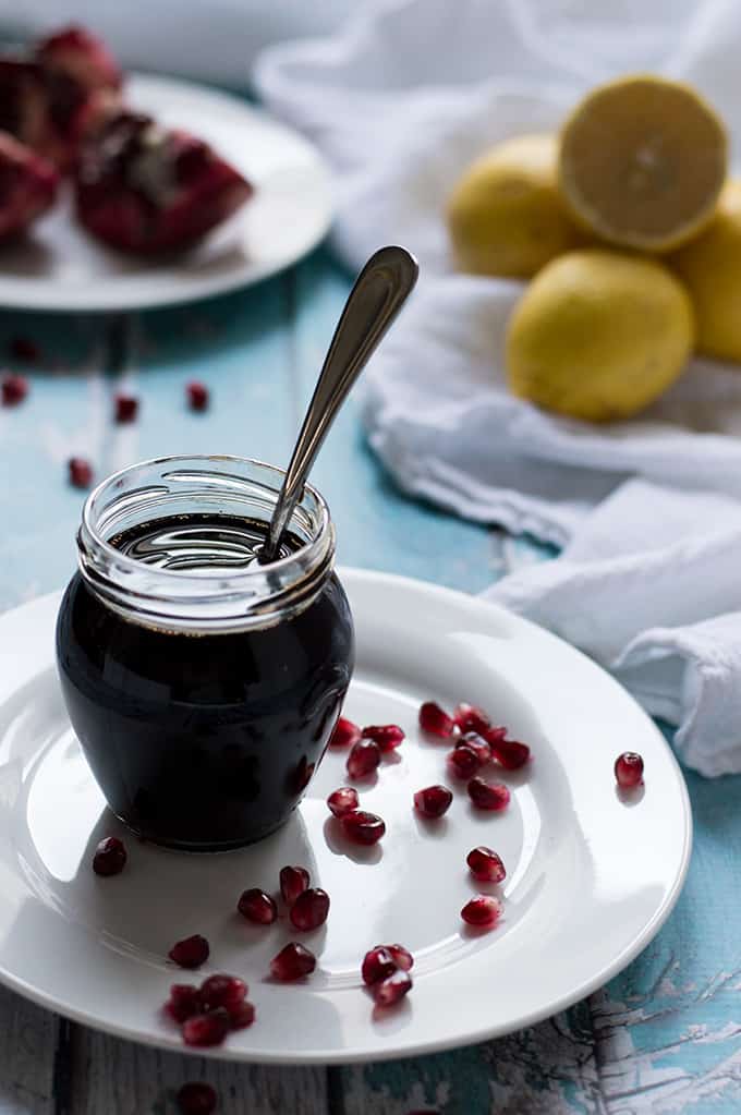 Glass jar of pomegranate molasses with a spoon inside and lemons and pomegranates in the background.