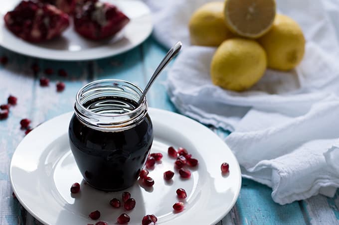 A jar of pomegranate molasses with a spoon inside and lemons and pomegranates in the background.