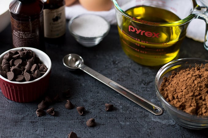 Bowls of chocolate chips, sugar, cocoa powder, and olive oil with a measuring spoon, bottle of vanilla extract, and a whole egg in the background.