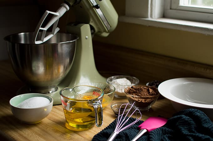 A stand mixer, sugar, cocoa powder, and olive oil, a whisk, silicone spatula, and ceramic bowl on a countertop.