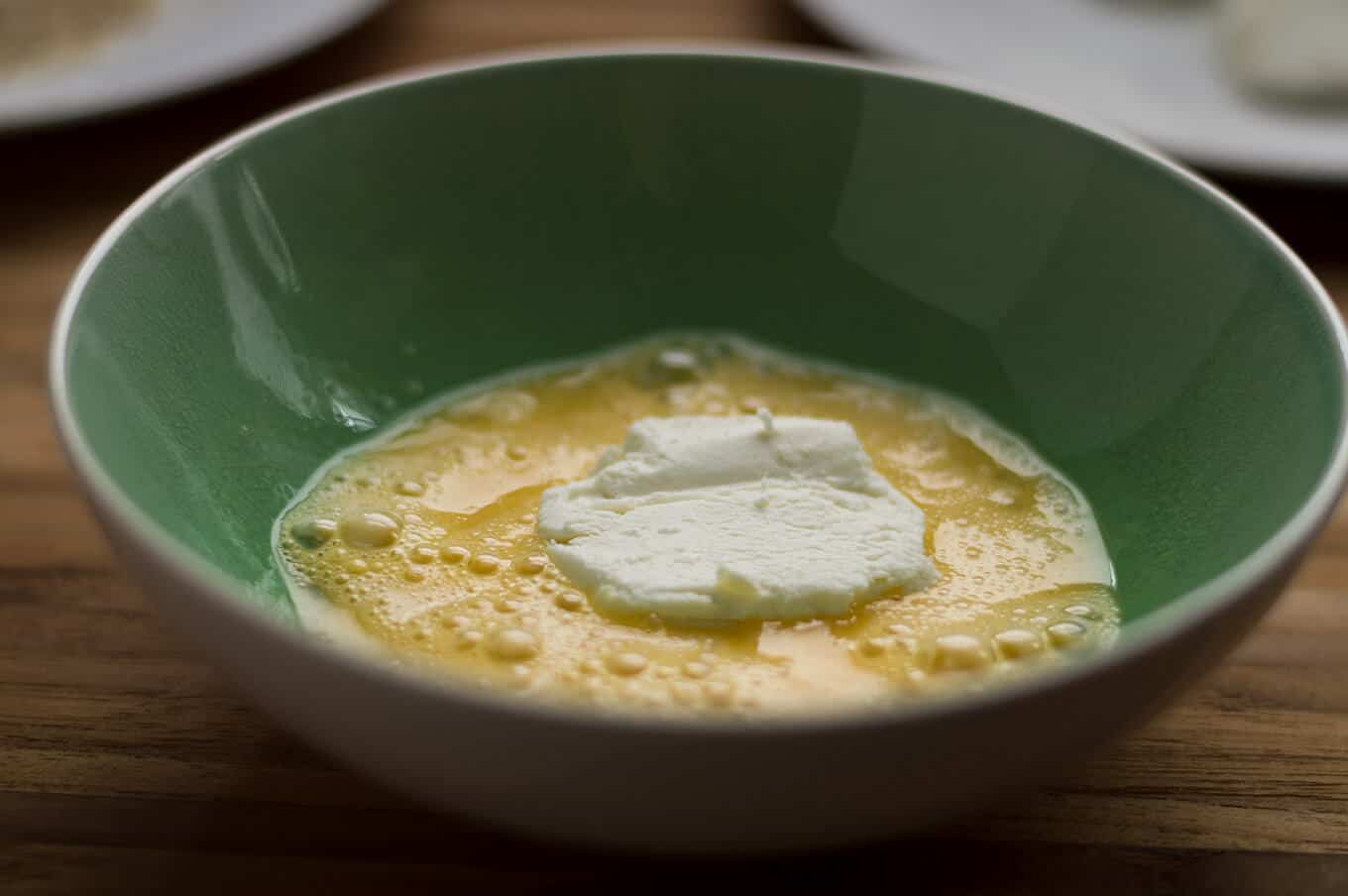 A close up a slice of goat cheese in a bowl of egg wash.