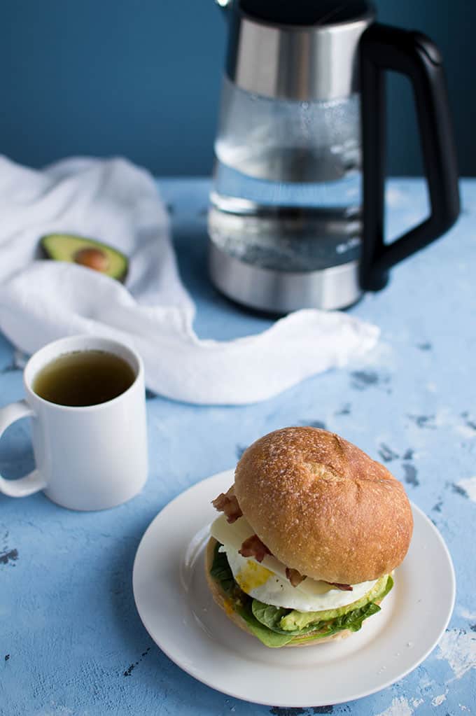 A mug of tea and breakfast sandwich on a table with a tea kettle in the background.
