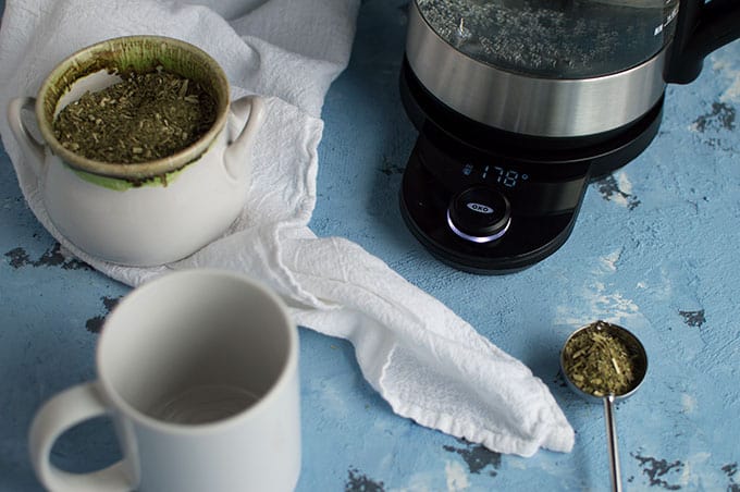 A tea kettle, empty mug, and scooped tea leaves.